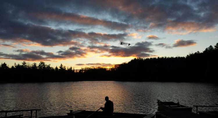 Beautiful sun set view of a man sitting by the lake. Clouds and sunset hue of orange and yellow can be seen in the sky.