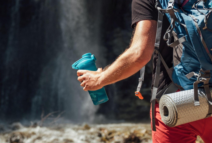 A man wearing a backpack on a hike near a waterfall holds a large blue water bottle.