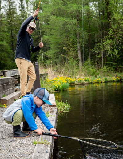 An adult male in a cap and sunglasses stands on a wooden fishing platform guiding a young boy in a blue shirt who is crouching at the water's edge.