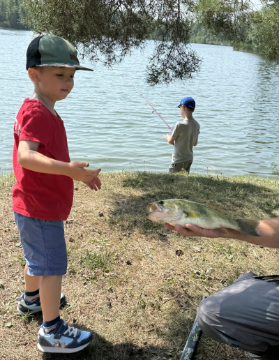 A young boy in a red t-shirt and green cap looking down at a fish on the grassy bank, while another child casts a line into the lake in the background.