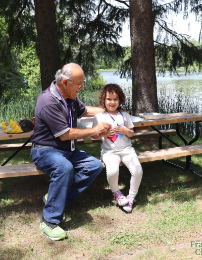 An older man and a young child sitting together on a wooden picnic table under the shade of tall pine trees near a peaceful lake.