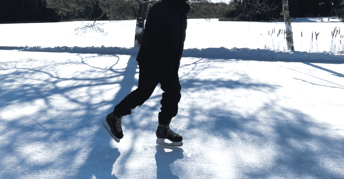 Franklin-Club-Winter-Pond-Skating A close-up action shot of a person's legs gliding on ice skates across a cleared natural ice rink on a bright, sunny winter day.