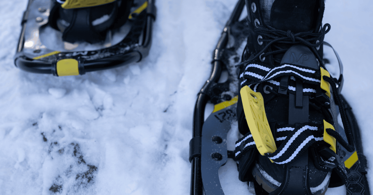Franklin-Club-Winter-Snowshoes-Gear A close-up view of a pair of modern black snowshoes strapped onto boots, resting on a field of fresh, white snow.