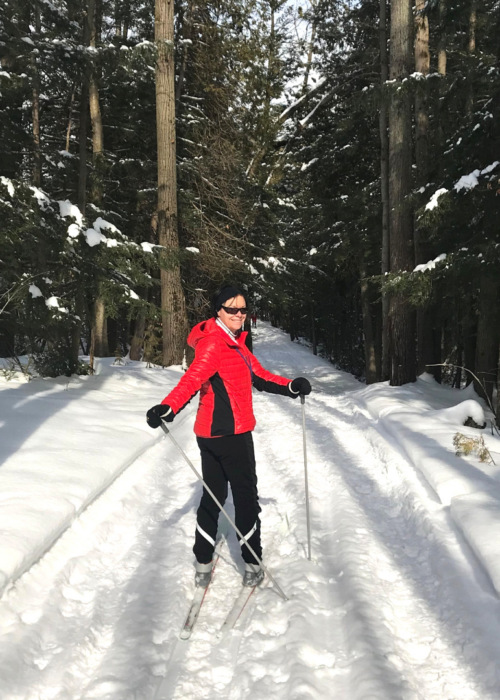 A woman wearing a vibrant red winter jacket and sunglasses cross-country skiing along a snow-covered trail slicing through a dense pine forest.