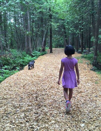 A young girl wearing a pink dress walking away from the camera down a wood-chipped path winding through a dense green forest.