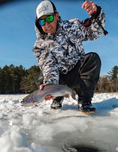 An angler wearing a winter camouflage jacket and sunglasses kneeling on a frozen lake, proudly presenting a large, freshly caught rainbow trout.