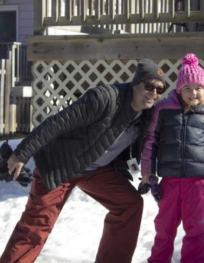 An adult and a young child dressed in warm winter snowsuits and hats laughing and playing together outside a rustic wood chalet in the fresh snow.