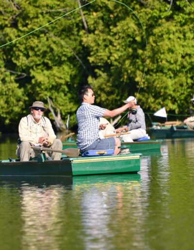 A group of club members enjoying a tranquil day, rowing and fishing from small green flat-bottomed boats on a scenic, tree-lined lake.