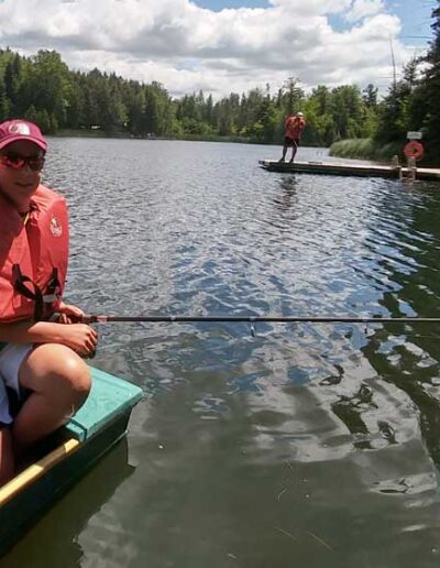 A close-up shot of a club member wearing a life jacket, seated in a classic green rowboat on a peaceful lake with a fishing line cast out.