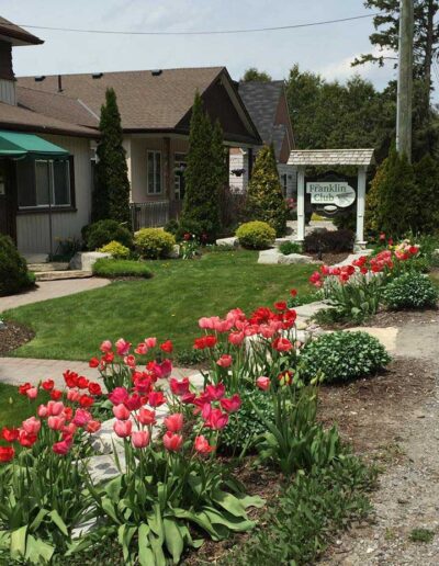 A beautifully landscaped front garden in bloom with bright red flowers, bordered by a manicured green lawn and a white wooden club sign in the background.