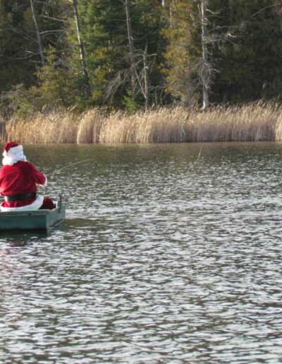 A festive scene of a club member wearing a bright red Santa hat rowing a small green boat on a calm lake with winter forest trees in the distance.