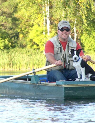 A club member in a red vest rowing a green wooden boat on a quiet lake with a small black-and-white dog sitting happily at the bow.