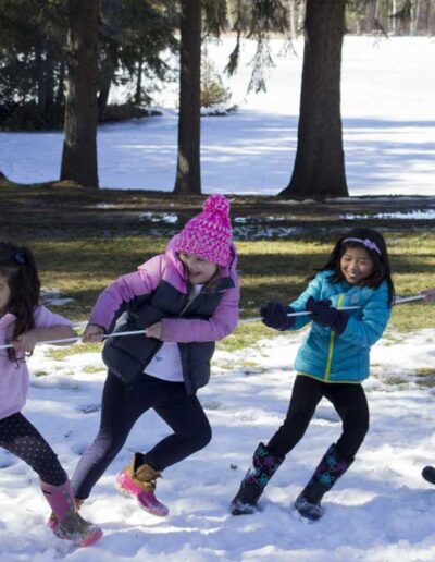 Four young children bundled up in colorful winter jackets pulling on a thick rope during an exciting game of tug-of-war on a snowy field.