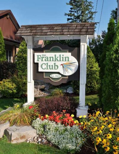 The beautiful white-arched Franklin Club entrance sign surrounded by lush green lawns, manicured evergreen shrubs, and colorful summer flowerbeds.