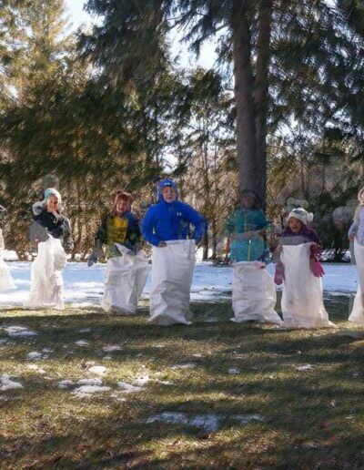 A group of participants of all ages lined up on a snowy lawn, jumping inside burlap sacks during a festive winter outdoor sack race.