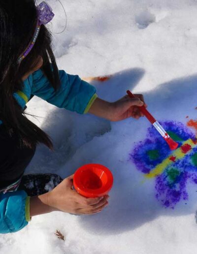 A child in a bright blue winter coat sitting in the snow, focused on painting or playing with colorful toys set on a small tray.