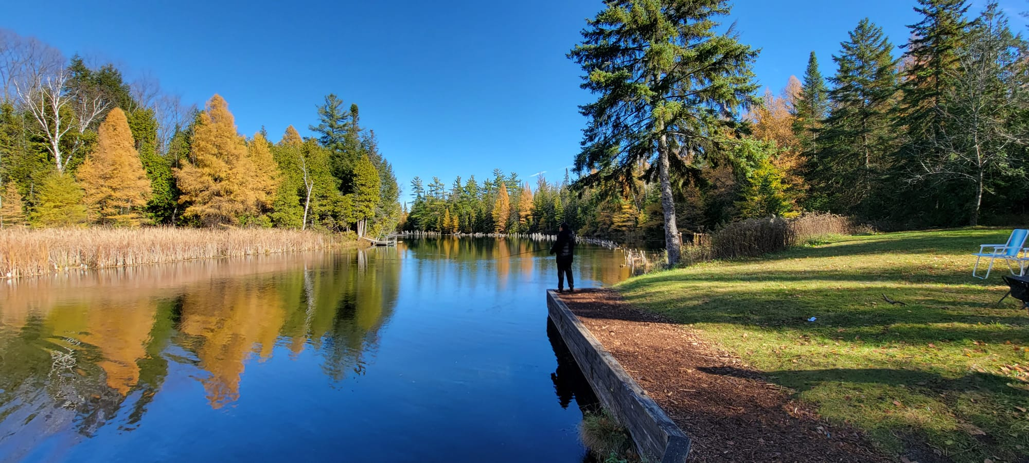 A wide, panoramic landscape photo by Bob Mok showcasing a calm lake with autumn foliage reflections and fly fishermen along the grassy banks.