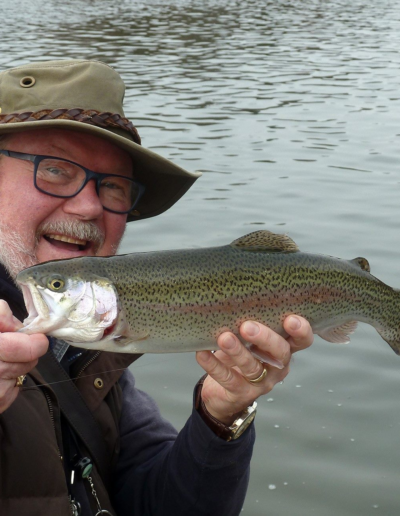 A close-up shot of a happy older man in a brown bucket hat and glasses holding a freshly caught rainbow trout close to the camera.