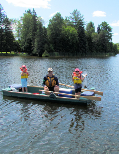 Two children wearing bright orange and yellow life jackets holding fishing rods while seated in a green rowboat on a sunny day.