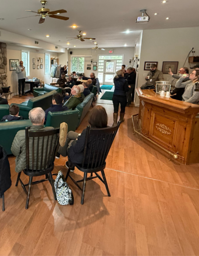 An indoor educational presentation inside the clubhouse lounge, with guests seated in armchairs listening to a speaker near a wooden podium.