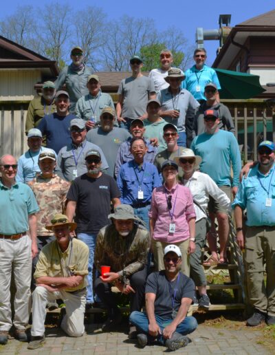 A large group of club members and guests posing for a group portrait on the wooden steps outside the main lodge on a sunny day.