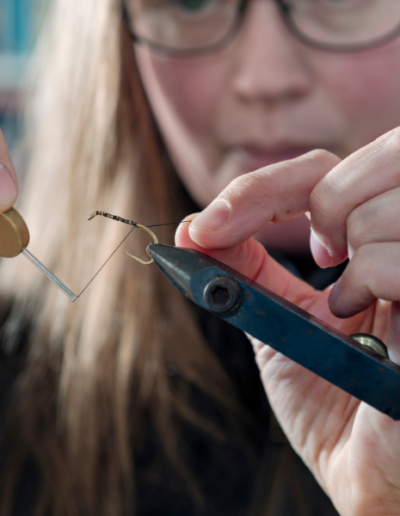 A close-up shot of a woman with glasses meticulously using a fine-tipped tool to thread and shape a delicate fly fishing lure.