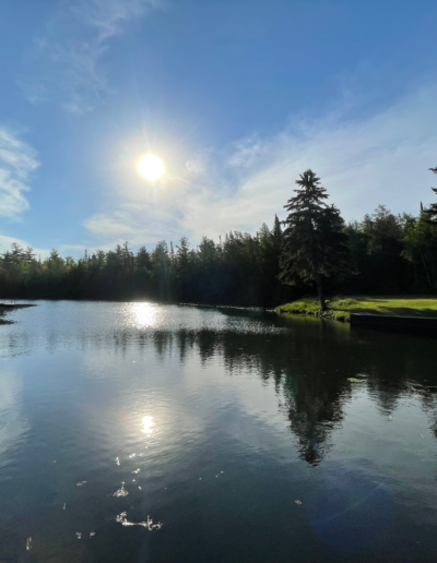 A scenic, wide-angle shot of a bright sunburst directly above a calm, dark lake framed by a dense forest line.