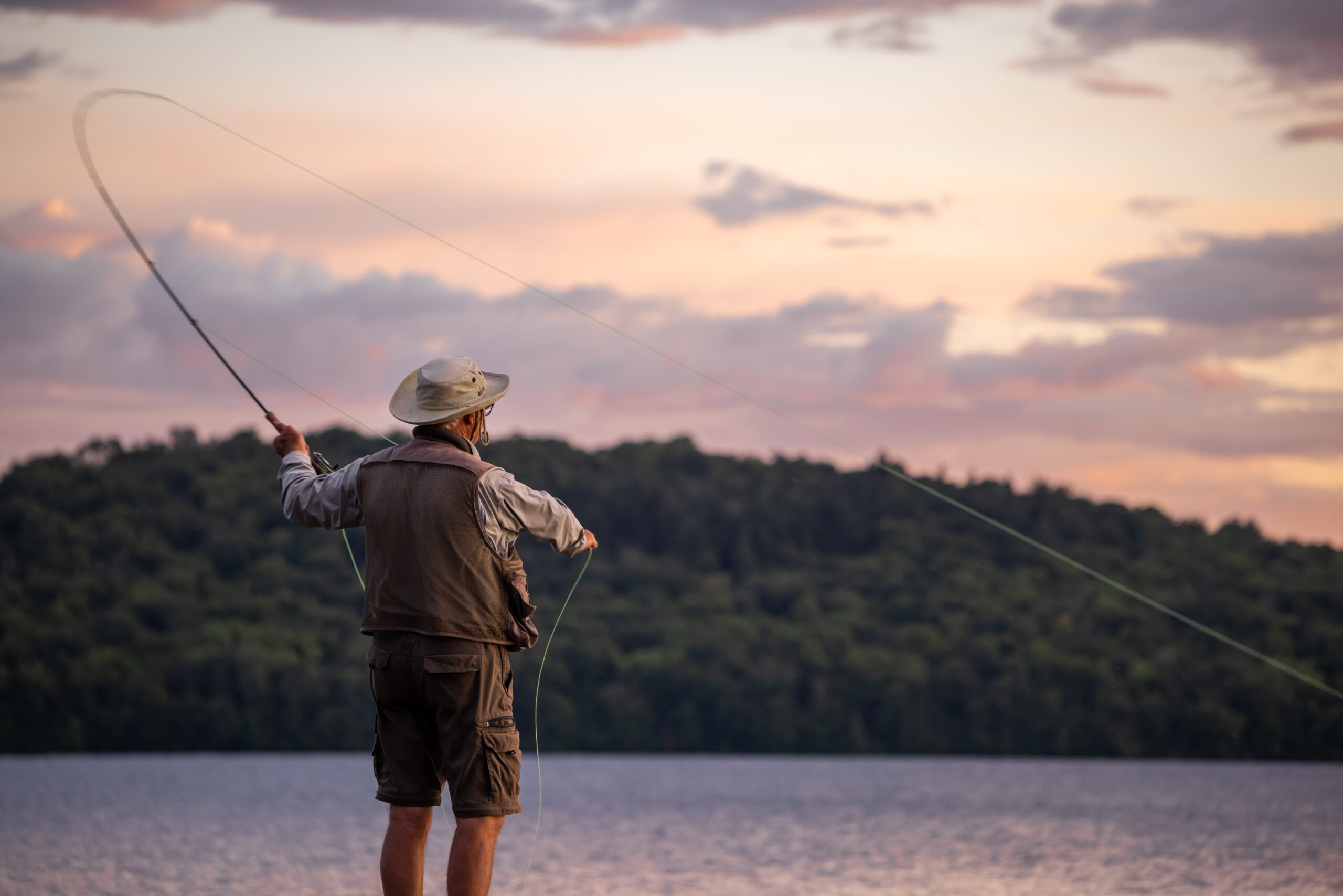 Franklin-Club-Senior-Angler-Sunset-Fly-Cast-WEBP A senior man wearing a sun hat fly fishing and casting his line over a calm lake at dusk, with the sky glowing in soft pastel sunset colors.