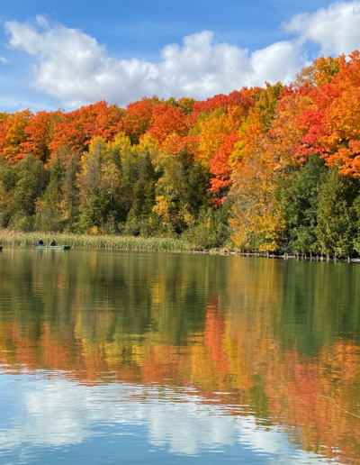 A beautiful lakeside landscape during peak autumn, featuring brilliant orange, red, and yellow trees reflecting perfectly in the calm water.