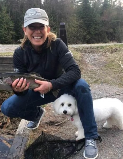 A smiling angler kneeling in a grassy field, proudly displaying a freshly caught dark fish next to a fluffy white dog.