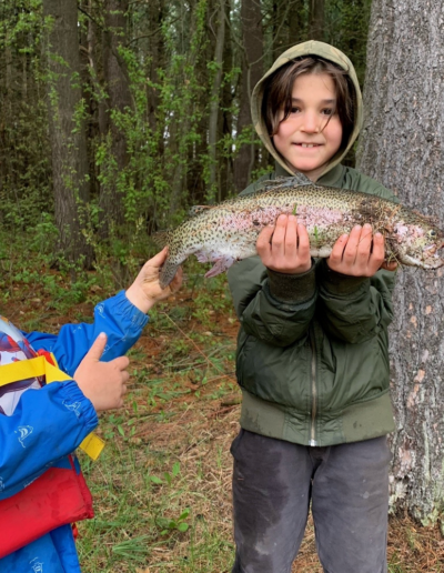 A young child wearing a green hooded jacket smiling while holding up a colorful trout, as a younger toddler looks on, with trees in the background.
