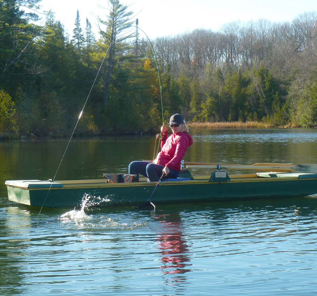 Franklin-Club-Solitary-Angler-Seated-Canoe-WEBP An angler in a pink jacket sitting relaxed in a green canoe on the calm water, with a dense green forest line in the background.