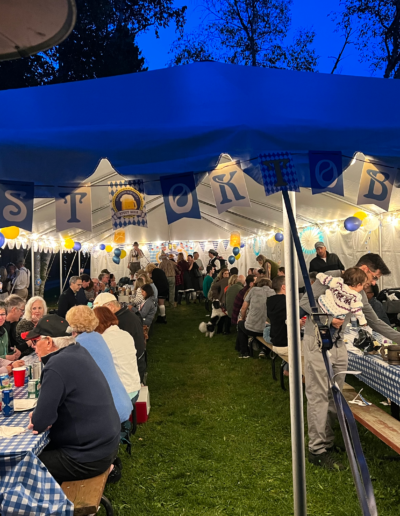 An evening outdoor gathering with guests dining at long banquet tables set up under a large, vibrant blue-lit canopy tent.