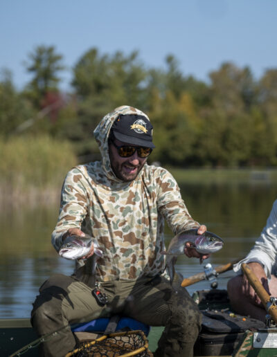 An angler proudly holding two freshly caught trout while seated next to his companion in a small green boat.