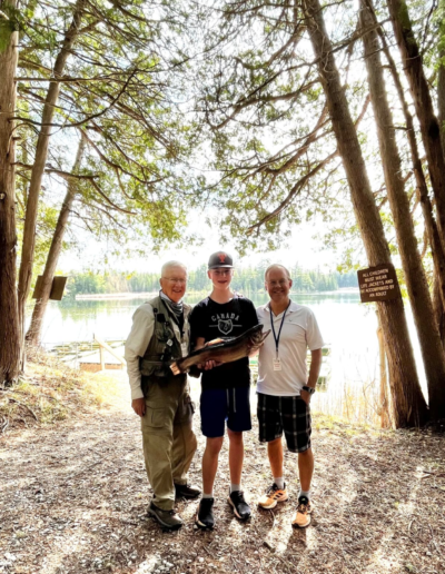 Three men standing together on the wooded shoreline of a calm lake under a canopy of trees.