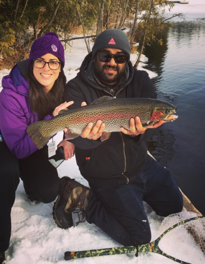 A smiling couple posing proudly by the water while holding up a large, vibrant rainbow trout they caught.