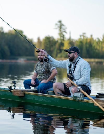 Two men enjoying a calm afternoon of fishing from a green canoe on a quiet, tree-lined lake.