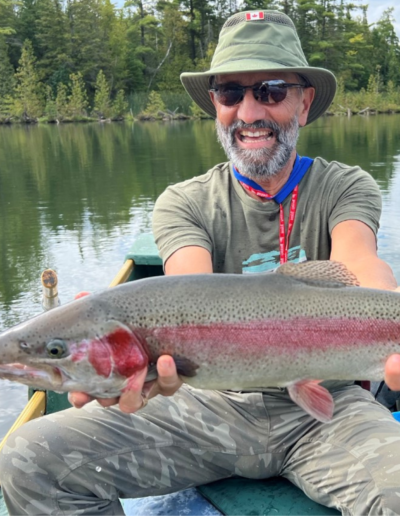 A smiling man wearing a sun hat and sunglasses holding up a massive, colorful rainbow trout on a bright day.