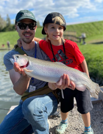 A man and a young boy in a red shirt smiling together while holding up a large rainbow trout they caught.