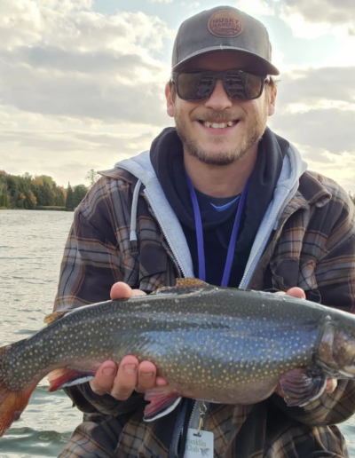 A smiling angler wearing a plaid shirt, cap, and sunglasses holding up a freshly caught trout over the water.