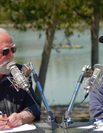 Two men sitting outdoors at a table with broadcast microphones, conducting an interview by a scenic lake.